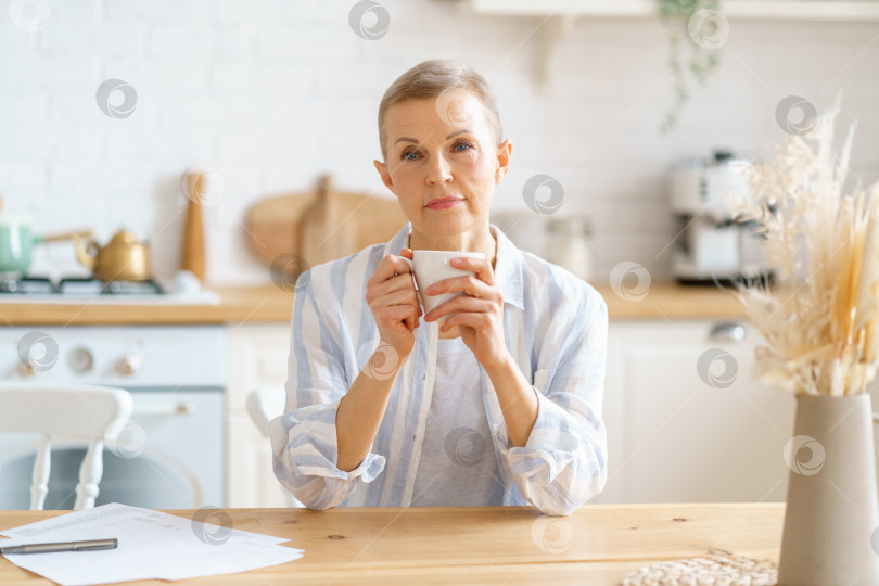 Скачать Привлекательная зрелая женщина начинает день с кофе или чая, сидя за деревянным столом на кухне фотосток Ozero