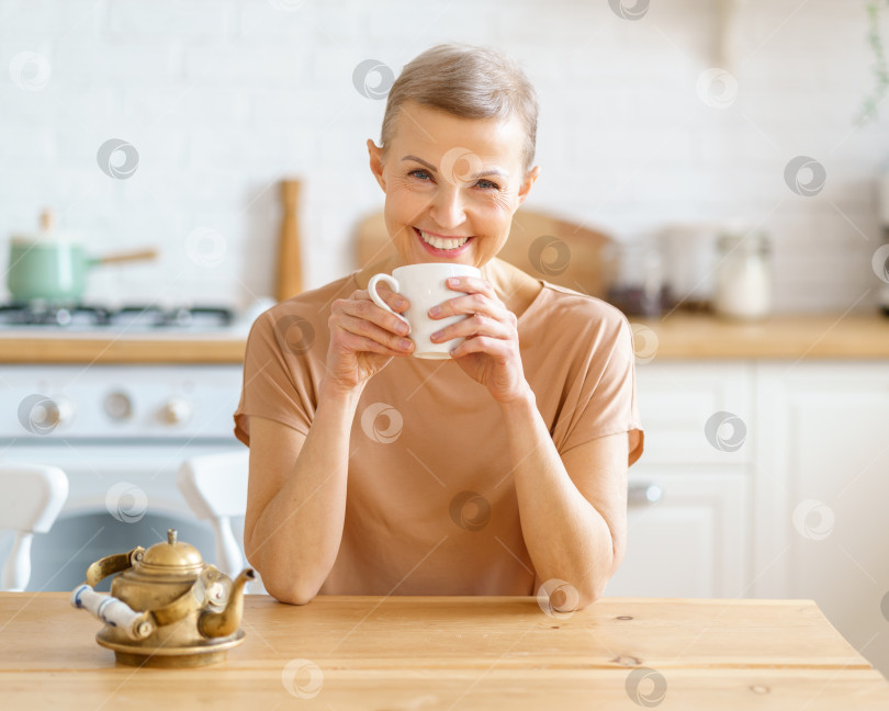 Скачать Привлекательная зрелая женщина начинает день с кофе, сидя за деревянным столом на кухне фотосток Ozero