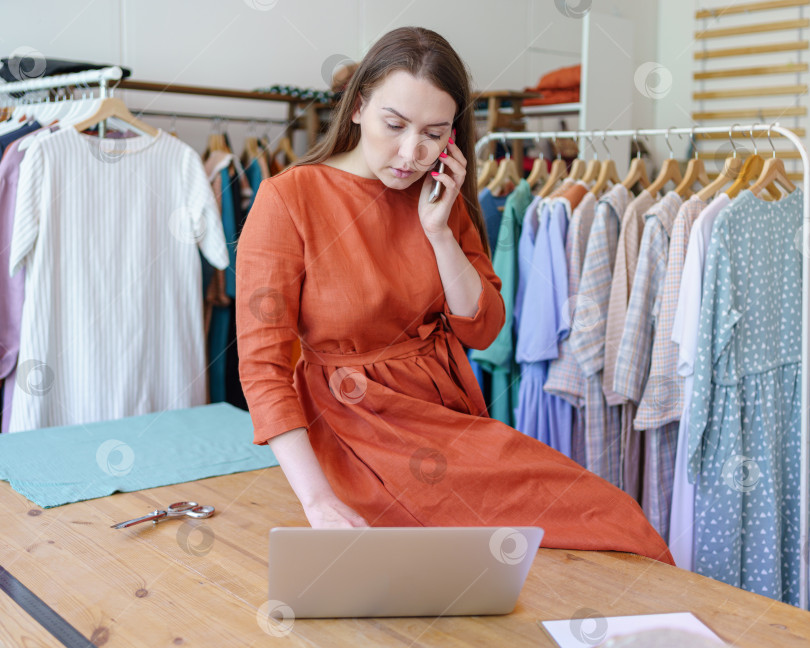 Скачать Женщина-модельер проверяет онлайн-заказ на ноутбуке, разговаривая с клиентом по мобильному телефону в своей мастерской фотосток Ozero