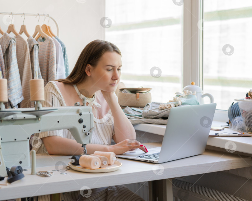 Скачать Женщина-модельер создает дизайн одежды онлайн, сидя на рабочем месте за швейной машинкой фотосток Ozero