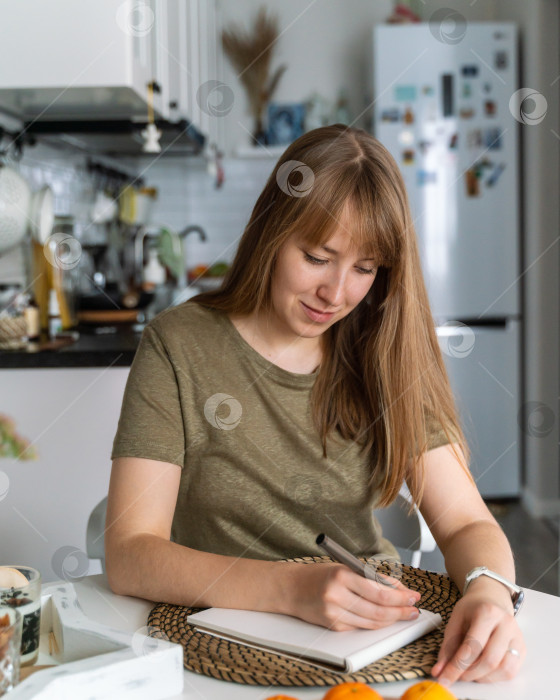 Скачать Молодая женщина делает пометки в блокноте, сидя на кухне. Планирует покупку. Составляет список продуктов фотосток Ozero