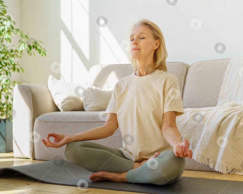 Скачать Пенсионерка медитирует и практикует йогу, сидя в позе лотоса на полу у себя дома фотосток Ozero