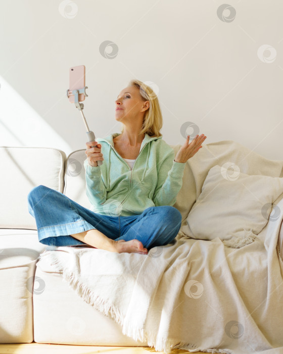 Скачать Позитивная пожилая женщина делает селфи на смартфон, отдыхая на диване у себя дома фотосток Ozero