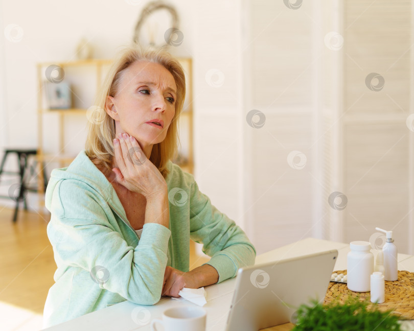 Скачать Пожилая женщина разговаривает онлайн с врачом о боли в горле, щитовидной железе, сидя дома на кухне фотосток Ozero