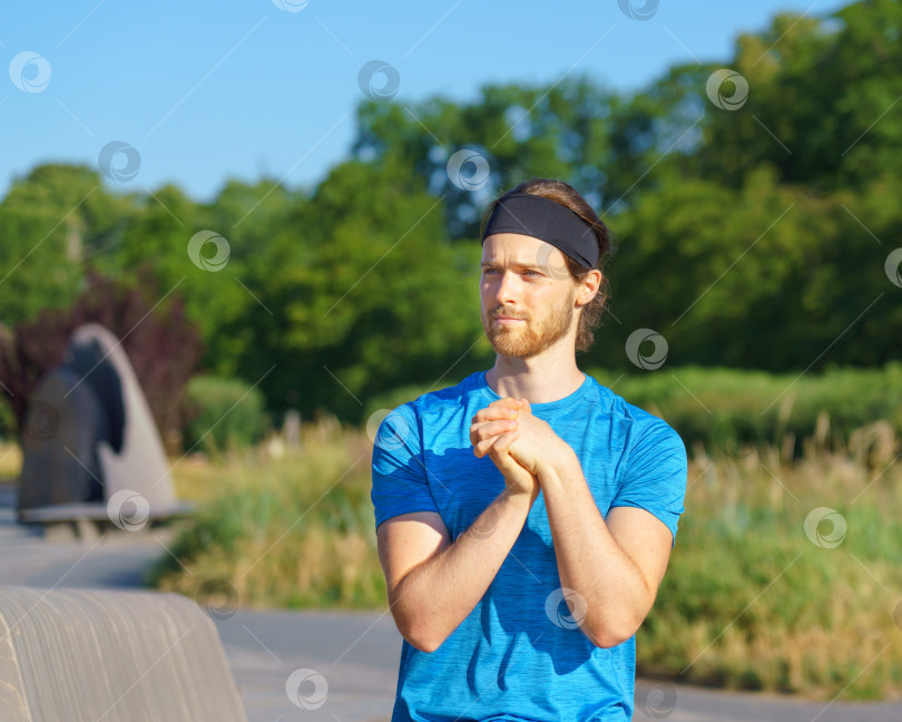 Скачать Молодой красивый подтянутый спортсмен в спортивной одежде смотрит в сторону, разминая руки и запястья на открытом воздухе фотосток Ozero
