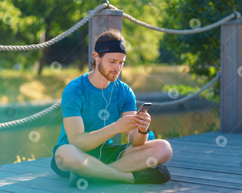 Скачать Спокойный молодой спортивный мужчина в наушниках пользуется современным смартфоном во время отдыха после тренировки в парке фотосток Ozero