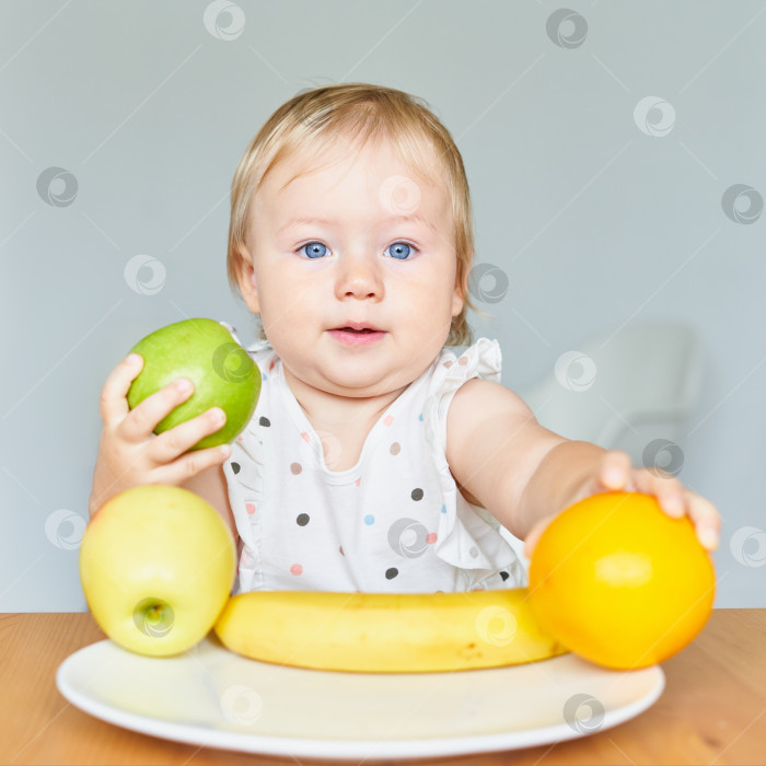 Скачать Очаровательная белокурая голубоглазая малышка держит в руках зеленое яблоко и апельсин. Тарелка с фруктами фотосток Ozero