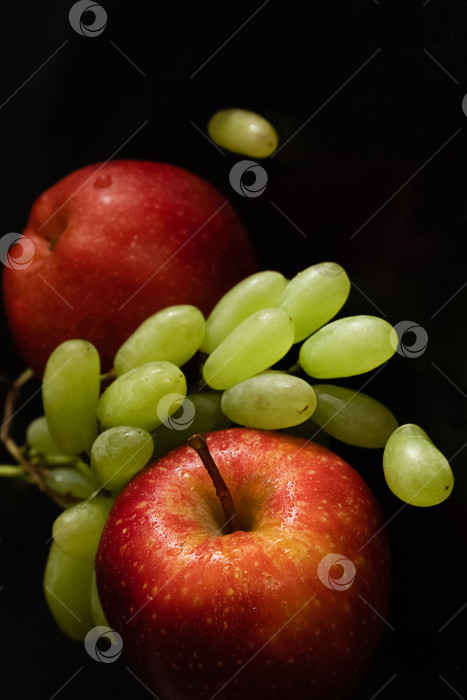 Скачать Два спелых свежих красных яблока с каплями воды на глянцевой кожуре и гроздь зеленого винограда на черном фоне фотосток Ozero
