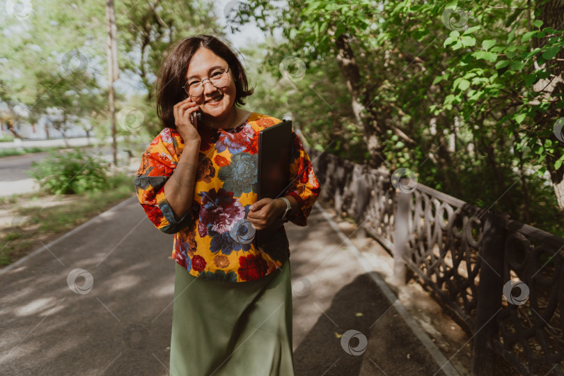 Скачать Веселая молодая женщина в очках разговаривает по мобильному телефону в пышном зеленом парке. фотосток Ozero