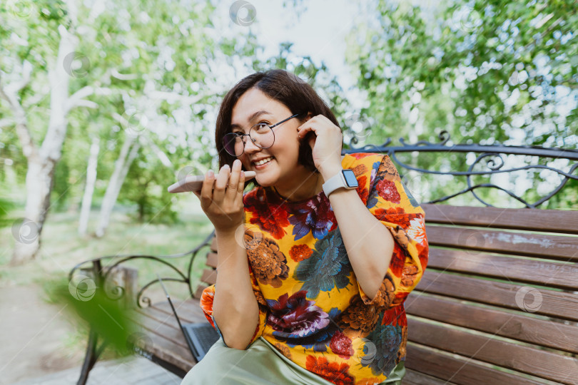 Скачать Счастливая женщина сидит на скамейке в зеленом парке, эмоционально разговаривая по телефону фотосток Ozero