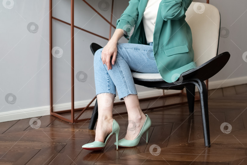 Скачать Женщина в синих джинсах и зеленой куртке сидит на стуле и хочет снять неудобные туфли на высоком каблуке фотосток Ozero