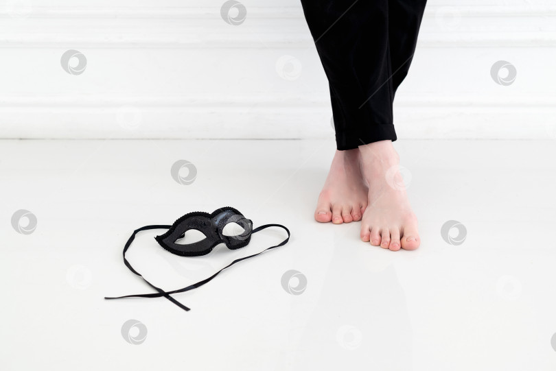 Скачать Черная карнавальная маска со шнурками лежит на белом полу возле босой ноги молодой женщины в черных брюках, которая стоит у белой стены фотосток Ozero