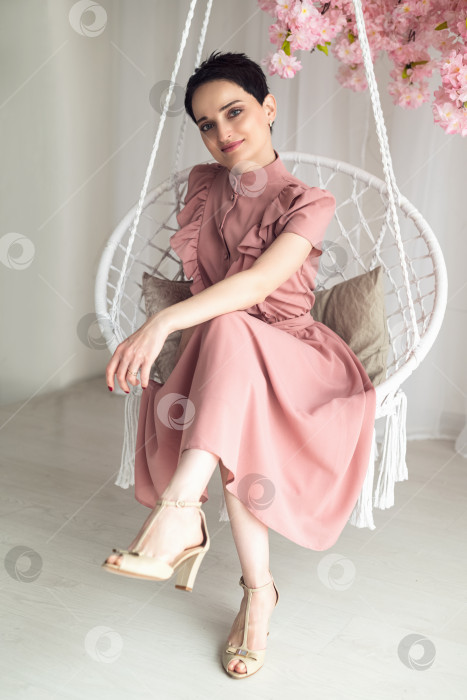 Скачать Молодая привлекательная брюнетка, одетая в розовое платье, сидит на декоративных плетеных качелях фотосток Ozero