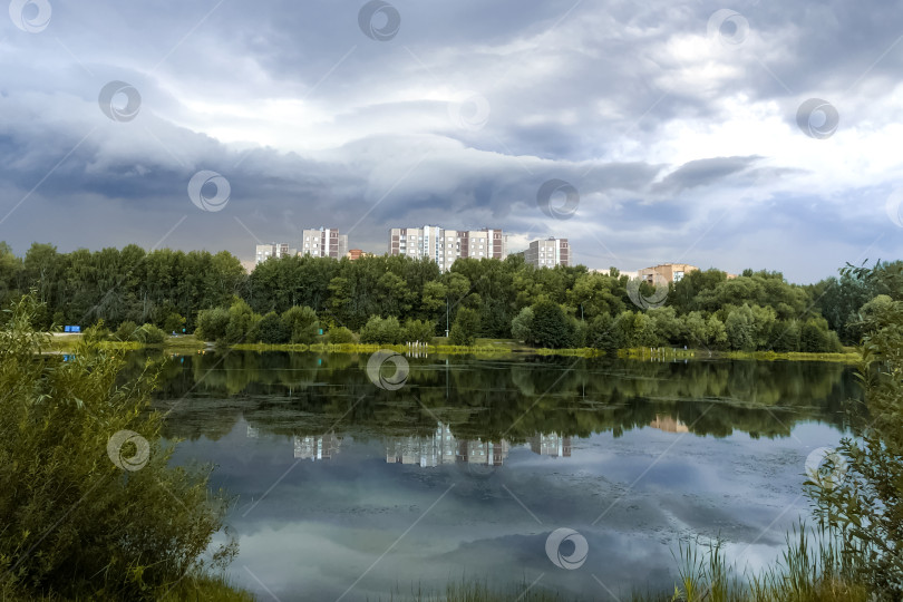 Скачать Летний городской пейзаж со спокойной водной гладью озера, облачным небом и отражающимися в воде многоэтажными жилыми домами фотосток Ozero