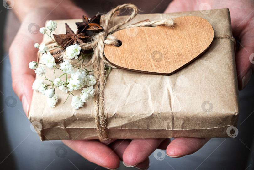 Скачать Подарочная коробка, обернутая в крафтовую бумагу, украшена деревянным сердечком и белыми цветами в человеческих руках. Празднование дня Святого Валентина фотосток Ozero