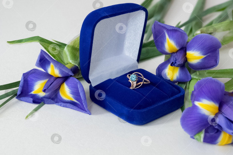 Скачать Обручальное кольцо из золота с голубым топазом в шкатулке для драгоценностей и букетом цветущих ирисов, разбросанных вокруг фотосток Ozero