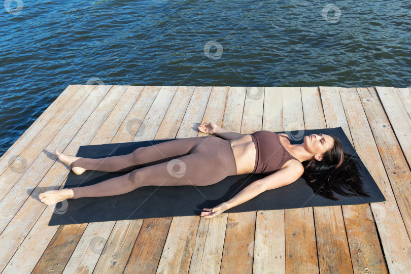 Скачать Женщина, лежащая на деревянной платформе на берегу озера, выполняет расслабляющее упражнение шавасана, занимается йогой в спортивной облегающей одежде на коврике фотосток Ozero