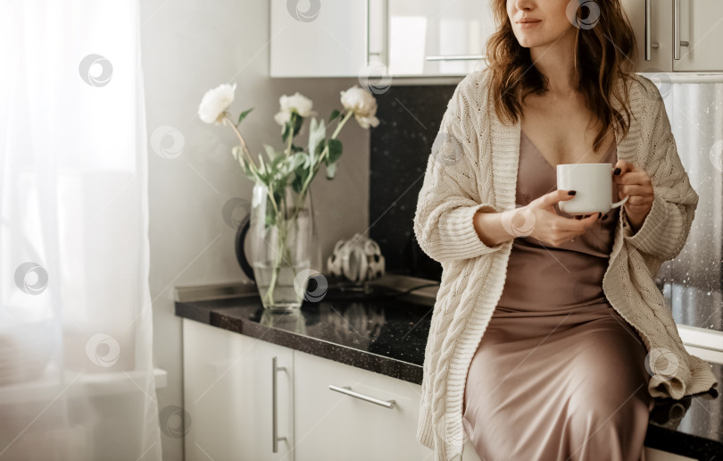 Скачать Молодая привлекательная женщина сидит на столе с чашкой горячего кофе в кухонной комнате с белым современным интерьером фотосток Ozero