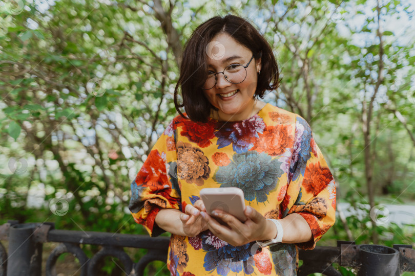 Скачать Счастливая азиатка в очках со смартфоном в летнем парке фотосток Ozero