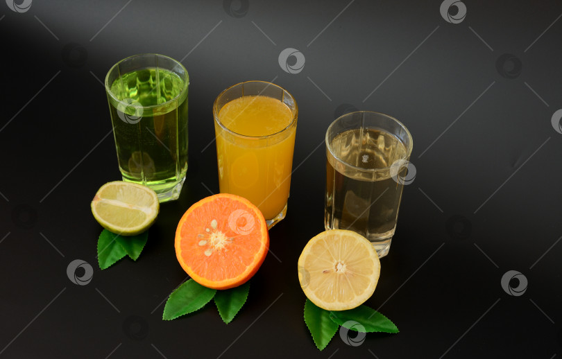 Скачать Три бокала с разными цитрусовыми соками стоят в ряд на черном фоне, рядом с половинками лимона, апельсина и лайма с листьями. фотосток Ozero