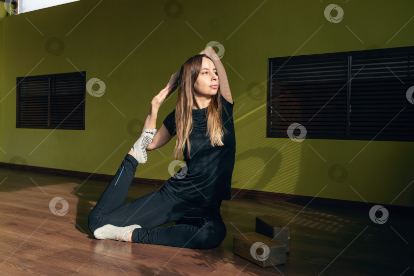 Скачать Привлекательная молодая женщина, выполняющая упражнение Эка Пада Раджакапотасана с захватом кольца, вариация позы голубя, практикующая йогу в черной спортивной одежде, сидя на коврике в студии фотосток Ozero