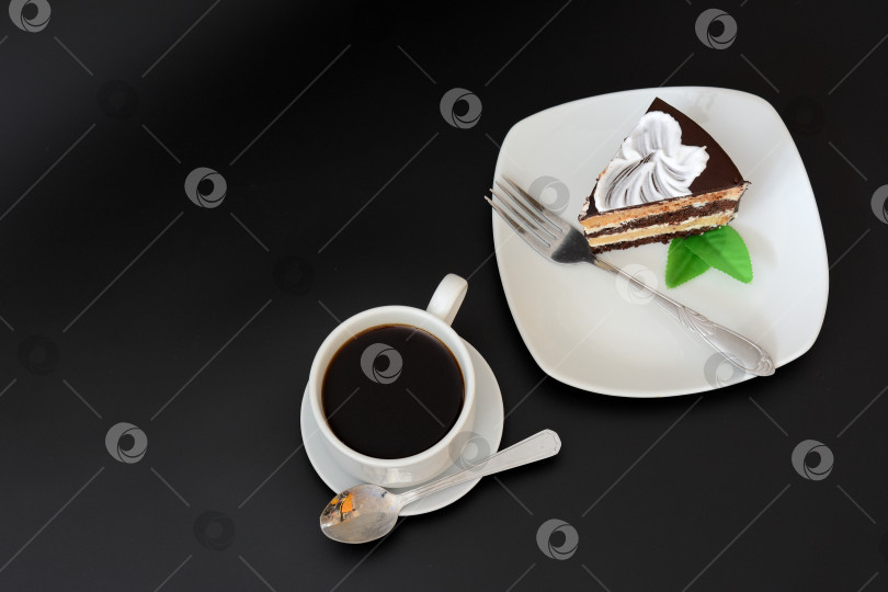 Скачать Белая керамическая тарелка с кусочком шоколадного бисквита с листьями мяты и вилкой на черном фоне, рядом с чашкой горячего черного кофе. фотосток Ozero