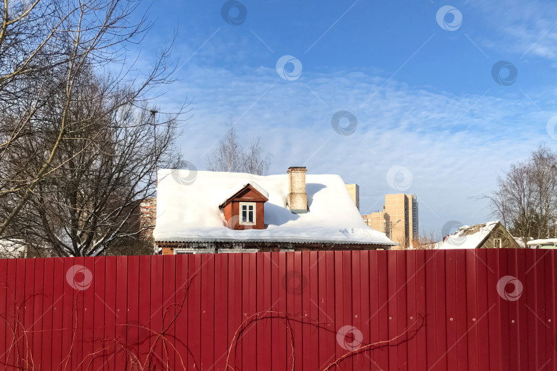 Скачать Заснеженная крыша деревенского дома видна из-за высокого красного забора фотосток Ozero