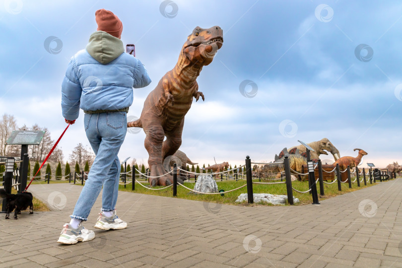 Скачать Девушка с собакой фотографирует фигуру тиранозавра в Динопарке Янтарного фотосток Ozero