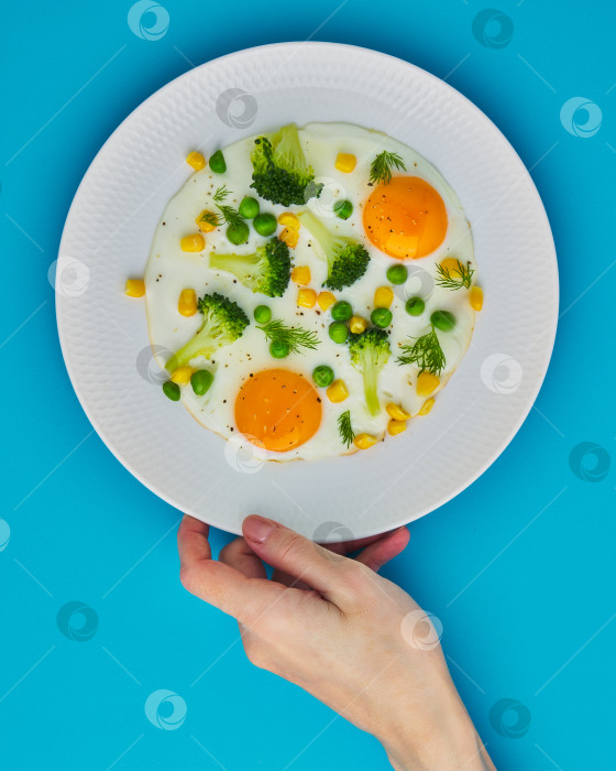 Скачать Женская рука касается тарелки с яичницей-глазуньей, брокколи, кукурузой на синем фоне фотосток Ozero