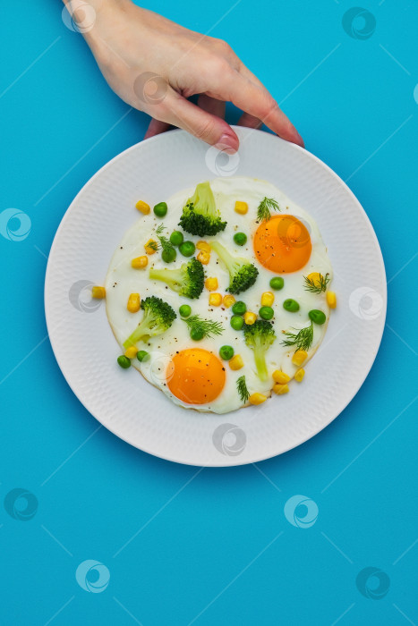 Скачать Женская рука касается тарелки с яичницей-глазуньей, брокколи, кукурузой на бирюзовом фоне фотосток Ozero
