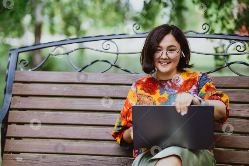 Скачать Улыбающаяся азиатка работает за своим ноутбуком, сидя на скамейке в парке фотосток Ozero