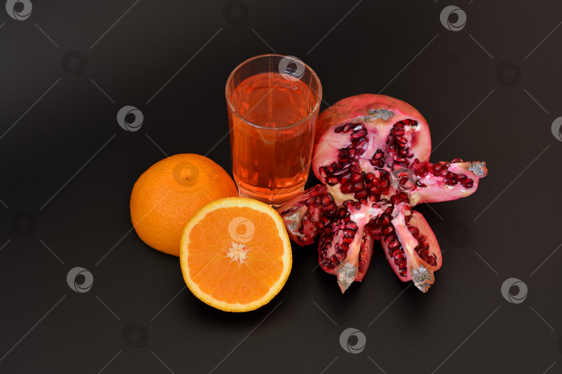 Скачать Высокий стакан фруктового сока на черном фоне, рядом с ним половинка спелого апельсина и разломанный плод граната с косточками. фотосток Ozero