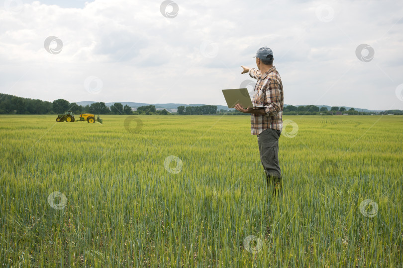 Скачать Мужчина-фермер с ноутбуком в поле с трактором, распыляющим пестициды на пшеничном поле. фотосток Ozero