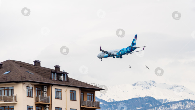 Скачать Boeing 737-800 авиакомпании NordStar Airlines пролетает над жилыми домами на фоне заснеженных вершин Кавказских гор. Крупный план. Шасси и закрылки самолета выпущены. фотосток Ozero