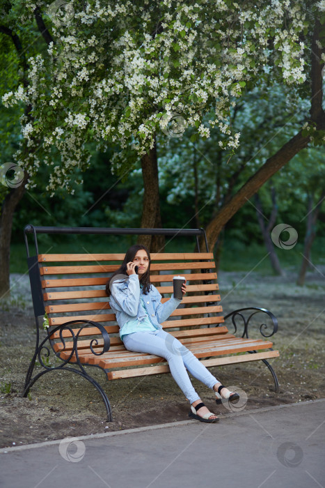 Скачать Девочка-подросток в джинсовой одежде разговаривает по телефону, сидя на скамейке в городском парке. фотосток Ozero