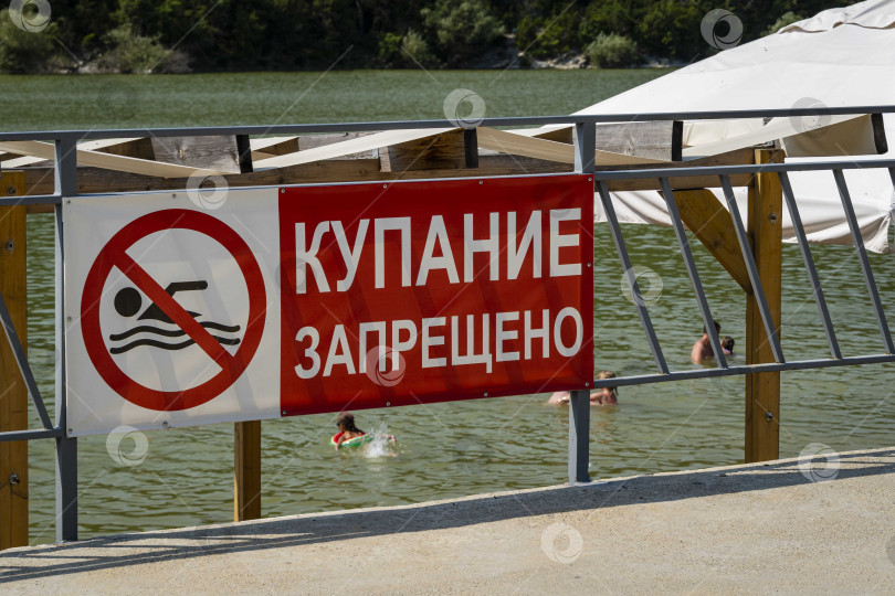 Скачать На оборудованном пляже с табличкой "Купаться запрещено" люди загорают и купаются. Кипарисовое озеро на живописном курорте Суко. Крупный план. Кавказские горы. фотосток Ozero