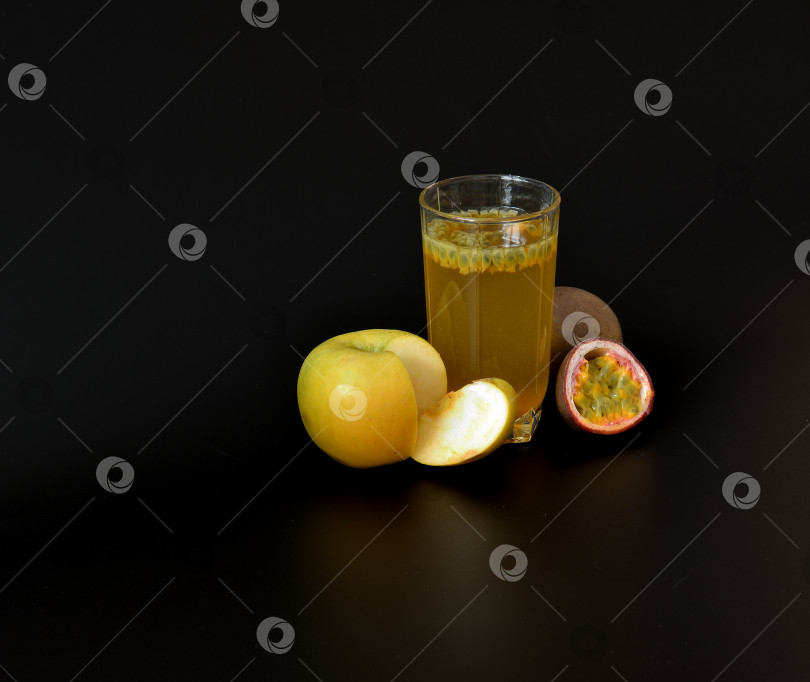 Скачать Стакан со смесью фруктовых соков на черном фоне, рядом с ним желтое яблоко и кусочки маракуйи. фотосток Ozero