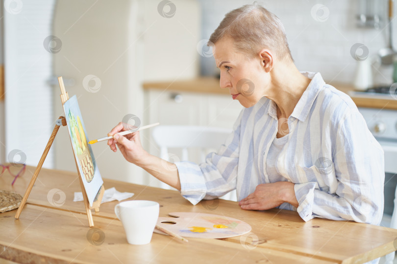 Скачать Творческая женщина средних лет рисует картину дома, сидя за деревянным столом на кухне фотосток Ozero