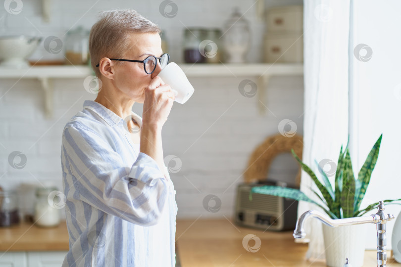 Скачать Привлекательная зрелая женщина начинает день с кофе, глядя в окно на кухне фотосток Ozero