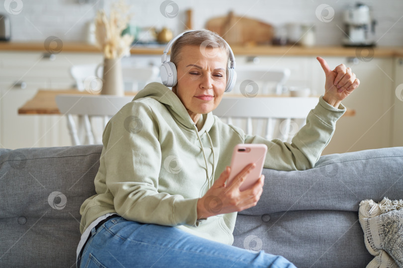 Скачать Счастливая пожилая женщина в наушниках слушает музыку на смартфоне, отдыхая дома на диване фотосток Ozero