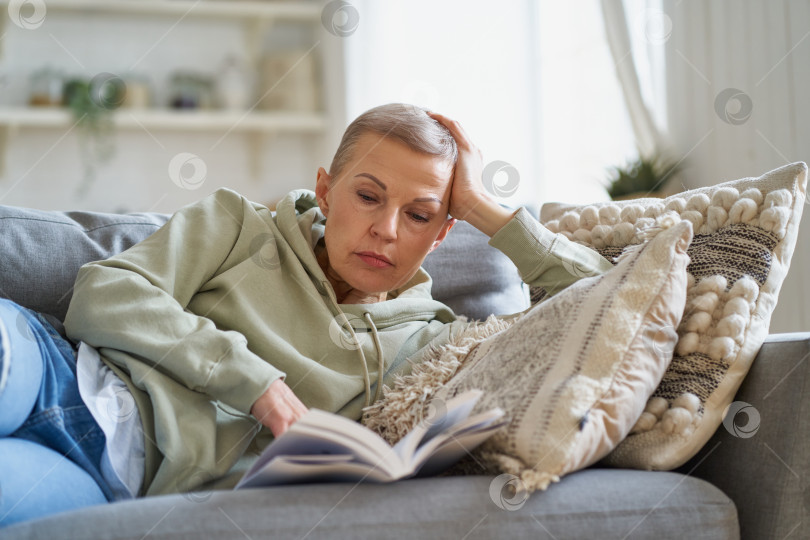 Скачать Веселая счастливая женщина читает книгу и улыбается, отдыхая на диване у себя дома фотосток Ozero