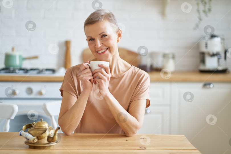 Скачать Привлекательная зрелая женщина начинает день с кофе, сидя за деревянным столом на кухне фотосток Ozero