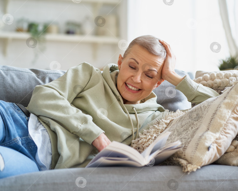 Скачать Веселая счастливая женщина читает книгу и улыбается, отдыхая на диване у себя дома фотосток Ozero