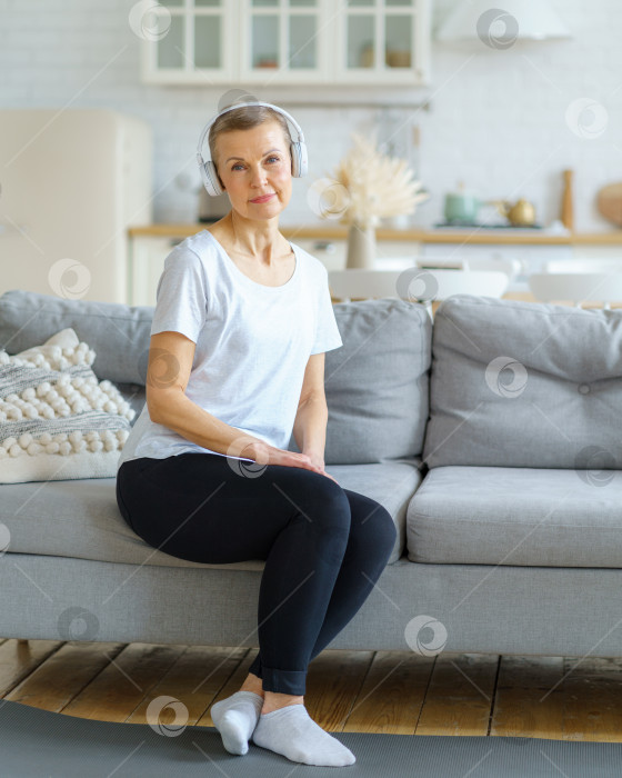 Скачать Портрет пожилой женщины, смотрящей в камеру, сидящей на диване в наушниках и слушающей музыку в гостиной фотосток Ozero