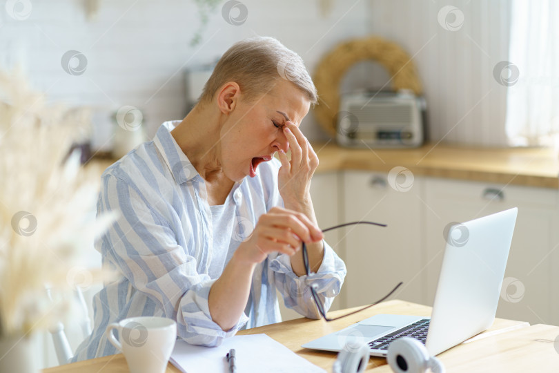 Скачать Женщина средних лет массирует нос и чувствует сонливость, работая на ноутбуке онлайн из дома фотосток Ozero