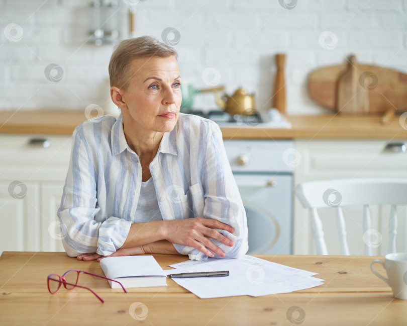 Скачать Задумчивая пожилая женщина, размышляющая о чем-то, сидя на кухне и работая с документами фотосток Ozero
