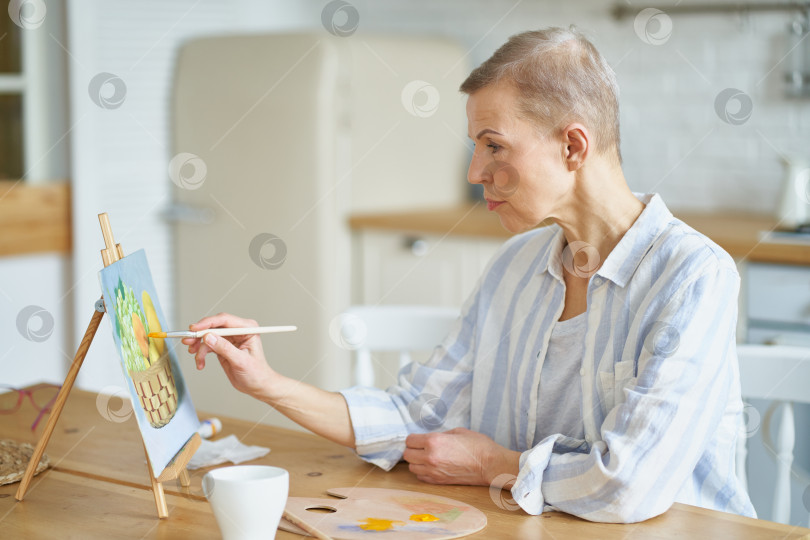 Скачать Творческая женщина средних лет рисует картину дома, сидя за деревянным столом на кухне фотосток Ozero