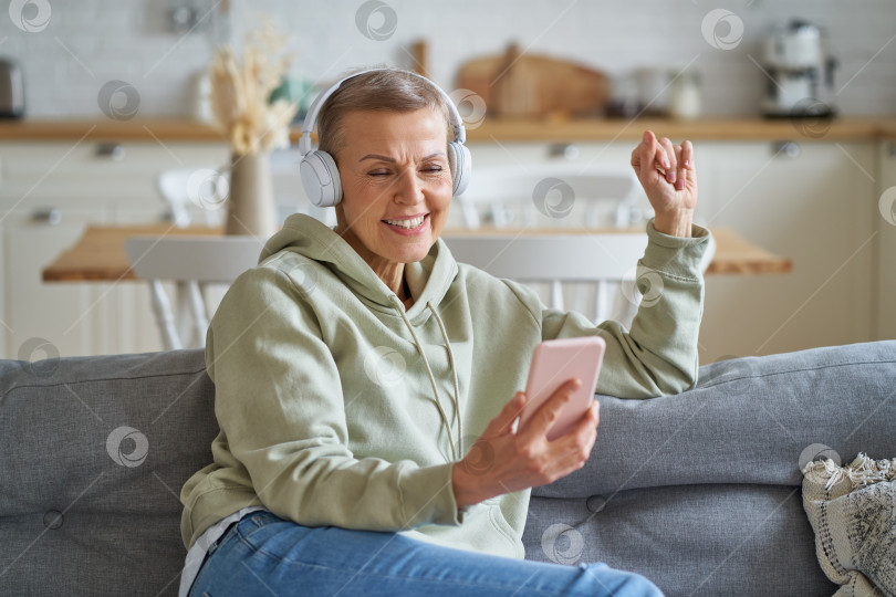 Скачать Счастливая пожилая женщина в наушниках слушает музыку на смартфоне, отдыхая дома на диване фотосток Ozero