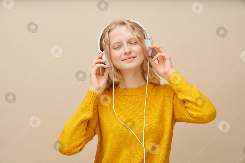 Скачать Крупным планом портрет молодой женщины с закрытыми глазами, слушающей музыку через наушники на бежевой стене. фотосток Ozero