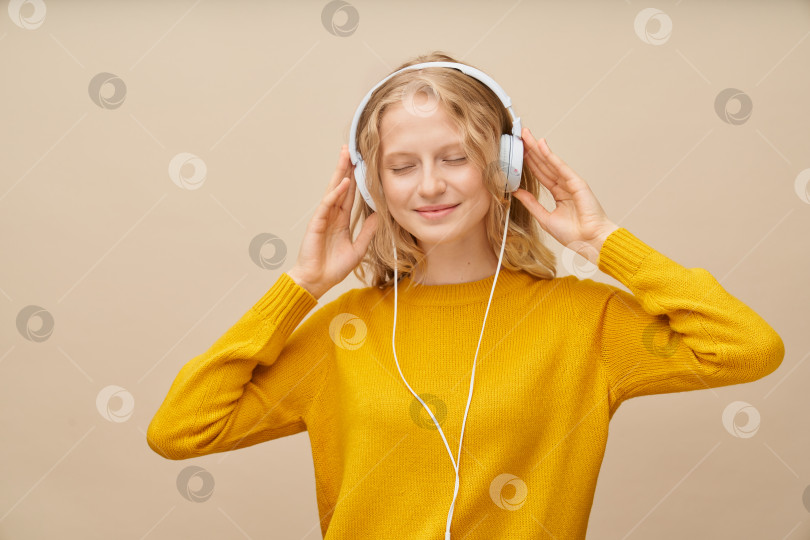 Скачать Крупным планом портрет молодой женщины с закрытыми глазами, слушающей музыку через наушники на бежевой стене. фотосток Ozero